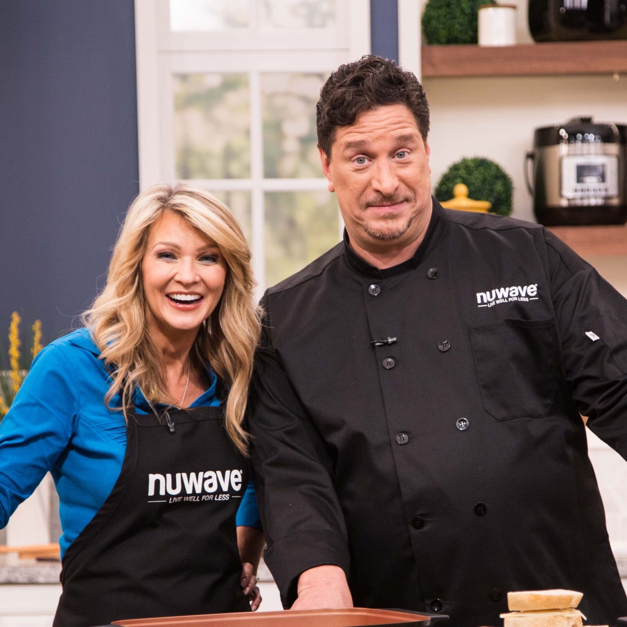 Two chefs posing in a kitchen, smiling and wearing aprons.